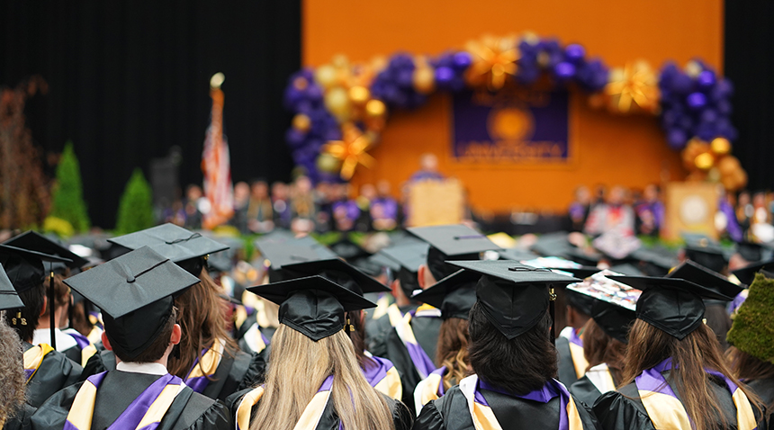 students seated at commencement facing the stage, in their caps and gowns